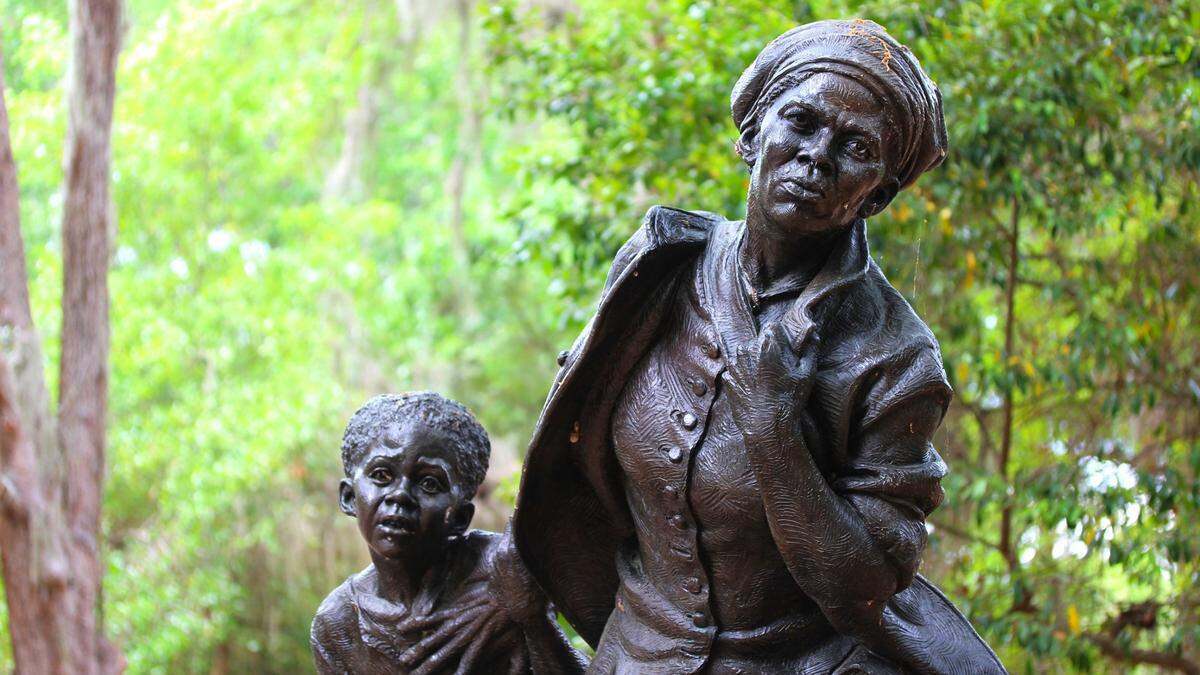 A bronze statue of Harriet Tubman shows the historical figure clenching her left, her eyes looking intently ahead with determination. Her left hand is placed on a fearful young African American girl's chest. 
