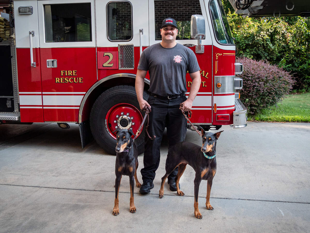 Lucas MacLellan, a firefighter with the Beaufort-Port Royal Fire Department, poses with Dobermans Ocho and Elke. The photo is one of 20 in a 2025 calendar that features local firefighters and therapy dogs. “It’s one-of-a-kind,” says Rebecca W. Bass, who took the photographs.