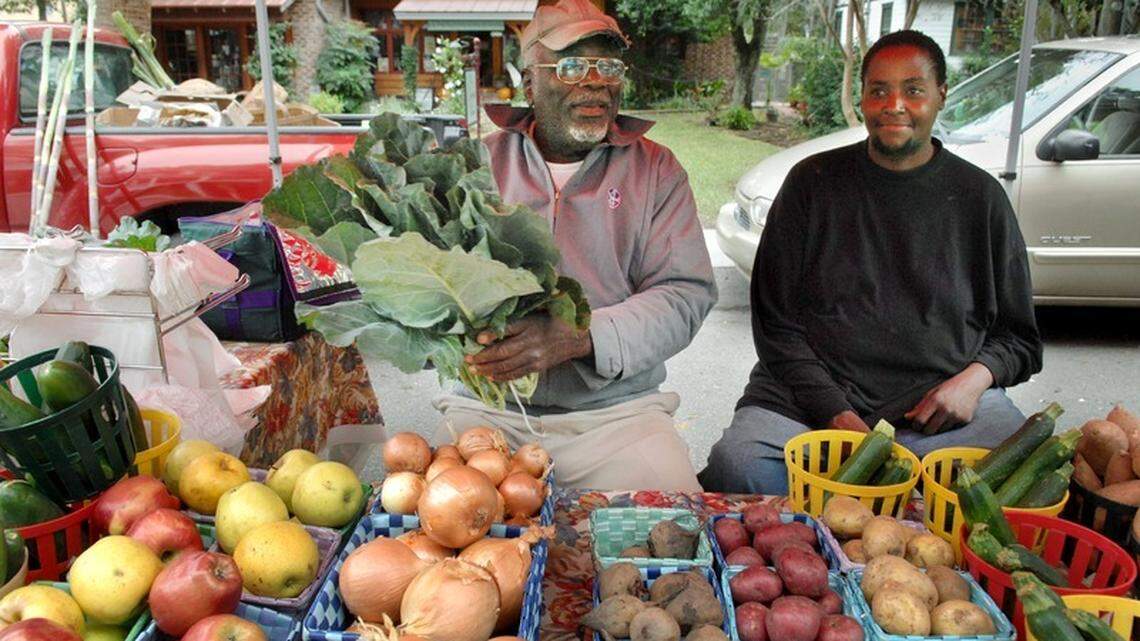 Donald D. Stevenson, of Stevenson Farm in Ridgeland, holds a bunch of collard greens as he sits under his display tent along with Eugene Habersham at the Farmers Market of Bluffton on Thursday. In December the market along Calhoun Street shifts to shorter winter hours.