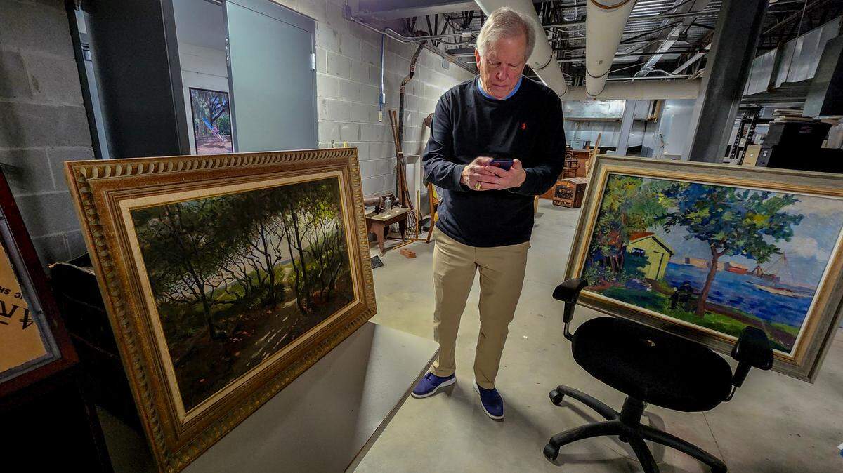John Warley, a Beaufort Art Museum board member, makes a phone call on Friday, March 1, 2024, to ask how an oil painting by Herbert Abrams, left, came into the museum’s hands held in storage space under Beaufort City Hall. While no one was sure, the untitled painting with dapples of light getting through a thick canopy of trees, will be one of a number of paintings, posters as well as a saber that will go to auction in the March 14 “Piece of Our Past Fundraising Auction” to be held in Beaufort.