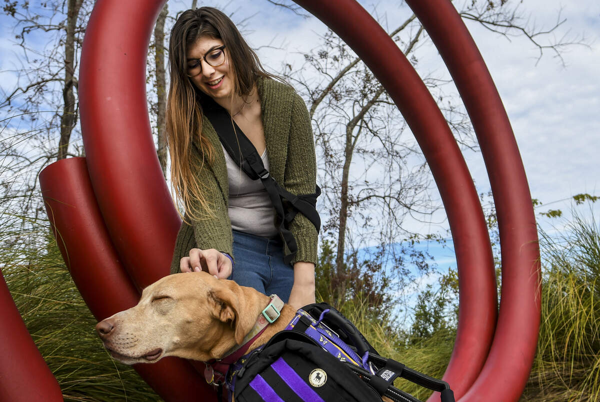 Serena Grumbine, 22, a Hilton Head Island resident, sits for a portrait at the island’s public sculpture “Carocol” at Shelter Cove Community Park on Monday morning as she scratches Millie’s head, her canine service animal. Last year, Grumbine had several surgeries to remove brain bleeds which led to months long therapies to help her speak and walk again at a center in California.