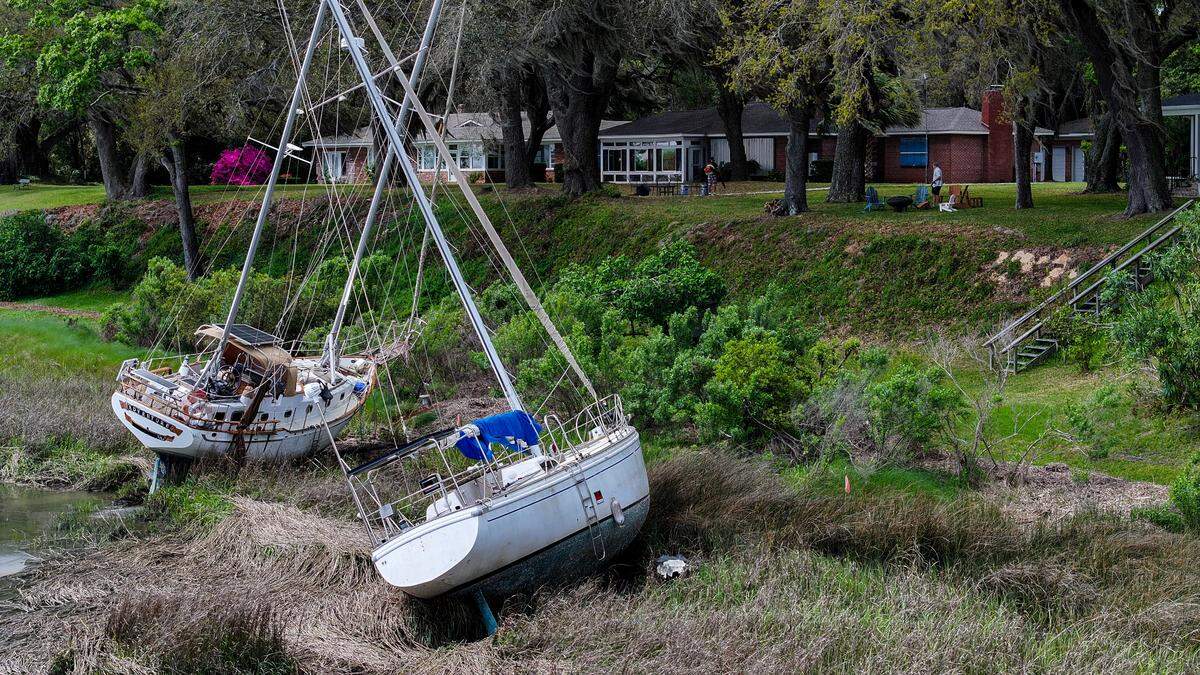 People work in the backyards of homes along Bay Street on April 3, 2025, as sailboats remain grounded on the bank of the Beaufort River in downtown Beaufort. In September, Tropical Storm Helene ripped numerous boats from their moorings and several, including these, remain.
