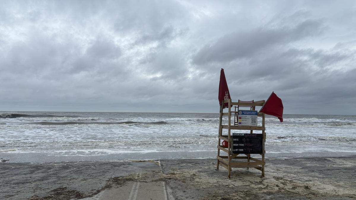 A lifeguard stand with a red flag sticking out from it sits on a grey, stormy beach.