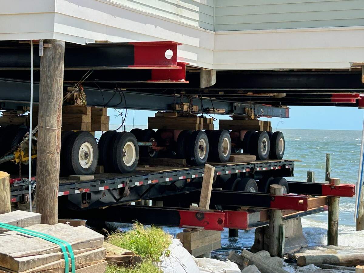 The white base of the home rests on top of steel and dollys, which is on top of a bridge in the water.