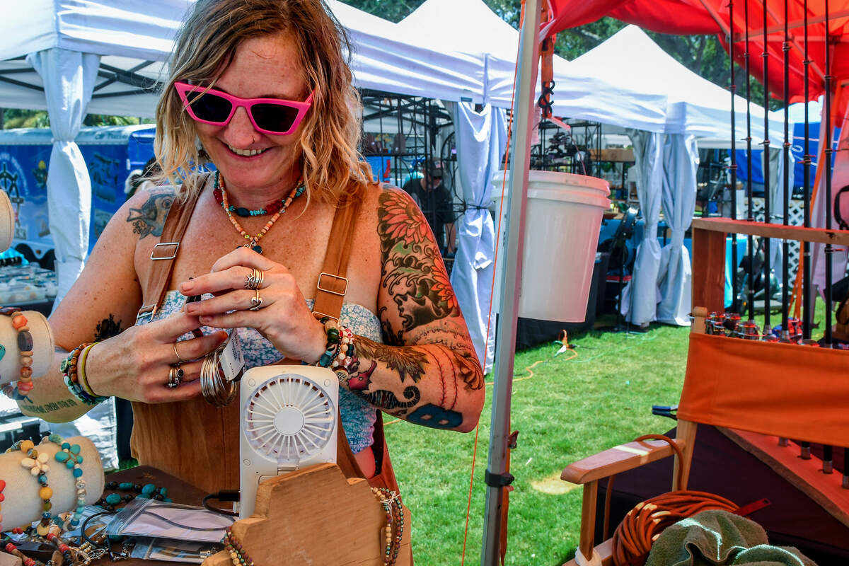 Charleston, S.C. jewelry maker Rebekah Aitken makes jewelry in front of her pint-sized fan on July 20, 2023 under her tent at Henry C. Chambers Waterfront Park at the Beaufort Water Festival. “I’d rather sweat than freeze,” Aitken said about such a small fan in such searing heat. “I don’t have room in my Jeep for anything bigger.”