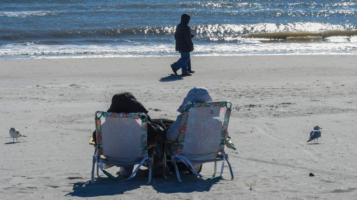 A couple wearing winter coats walks by a bundled duo in beach chairs as a stiff breeze comes off the Atlantic Ocean on the frigid morning of Tuesday, Jan. 7, 2025, on Hilton Head Island. For the first weeks of the new year a polar vortex from the high Arctic will bring freezing temperatures to parts of the deep south.