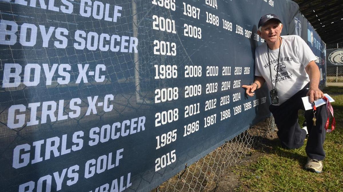 Hilton Head Island High School cross country coach Bill Wrightson poses on Wednesday with a banner listing boys and girls cross country championships. All of the championships, other than that of 1986, were won by teams he coached. Missing from the sign are boys state titles in 2014, 2015 and, of course, 2017. They also won it in 2009, and were second in 2008 and 2012.